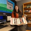 A woman with long dark hair and glasses stands in a digital preservation lab, holding open a scrapbook filled with old family photos. She wears a patterned dress with an orange cardigan and smiles at the camera. Behind her, a colorful banner reading 'Memory Lab' hangs on the wall, with a workstation featuring a computer, VHS tapes, and archival equipment visible.